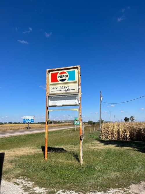 A weathered sign for "Six Mile Cafe" with a Pepsi logo, set against a clear blue sky and cornfield backdrop.