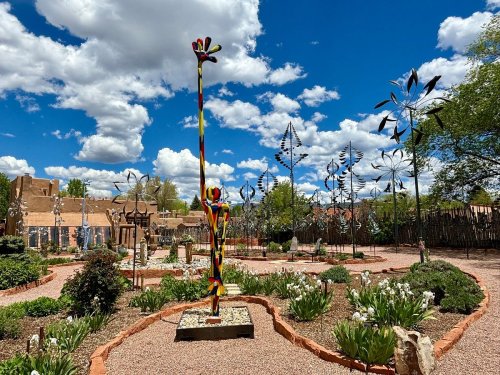 Colorful sculptures and wind spinners in a garden under a bright blue sky with fluffy clouds.