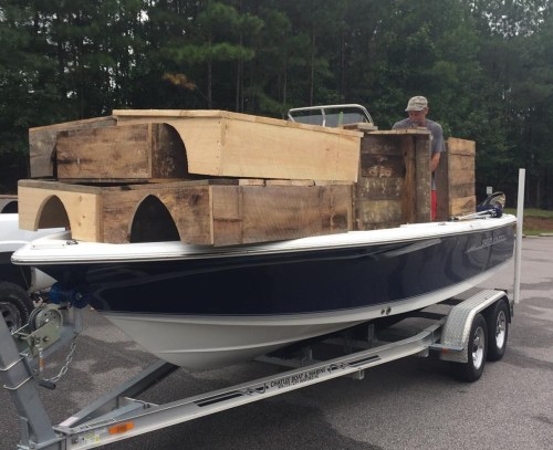 A man loading wooden crates onto a blue and white boat parked on a trailer in a wooded area.