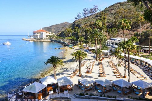 A scenic beach with palm trees, lounge chairs, and a view of a coastal resort and hills under a clear blue sky.