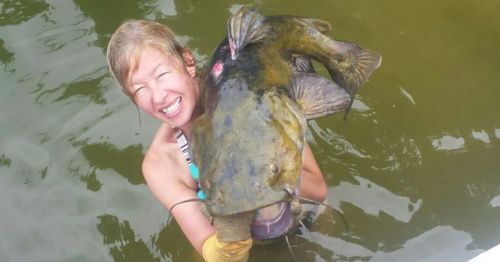 A smiling person in water holds a large catfish, showcasing their catch with excitement.