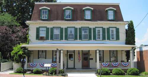 Historic two-story house with green shutters, a brown roof, and patriotic decorations on the porch.