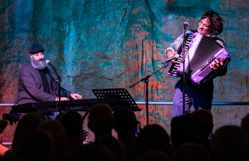 Two musicians perform on stage, one playing piano and the other playing accordion, with a colorful backdrop.