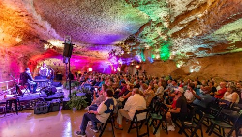 A lively concert in a colorful cave, with a large audience seated and performers on stage.