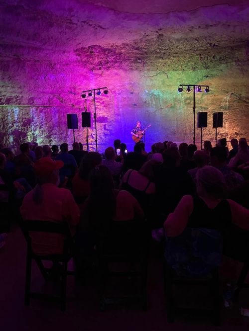 A musician performs on stage in a colorful cave, with an audience seated in front, illuminated by vibrant lights.