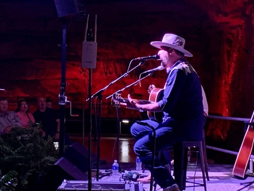 A musician performs on stage in a cave, with red lighting and an audience in the background.