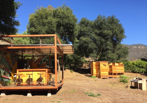 A wooden structure with a bar area and two storage units, surrounded by trees and mountains under a clear blue sky.