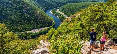 Two hikers stand on a rocky overlook, gazing at a winding river surrounded by lush green hills.