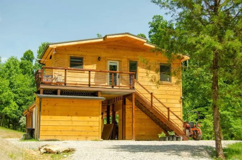A wooden two-story house with a balcony, surrounded by trees and a gravel driveway.
