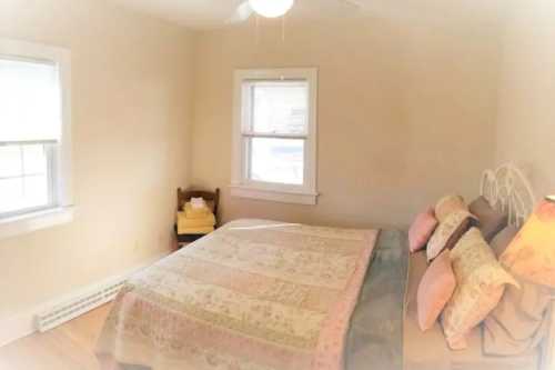 Cozy bedroom with a large bed, pastel bedding, and natural light from two windows. A chair with towels is in the corner.