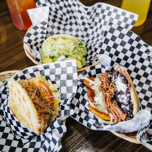 Three tacos in checkered paper baskets, featuring shredded meat, toppings, and a green dish in the background.