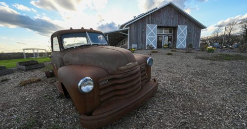 An old, rusty truck in front of a rustic barn with a cloudy sky in the background.