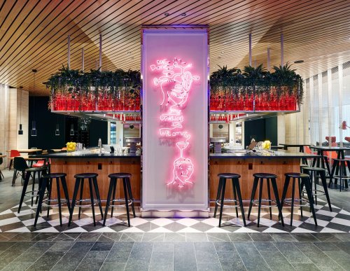 A modern bar with a neon sign, surrounded by plants and stools, featuring a stylish interior design.