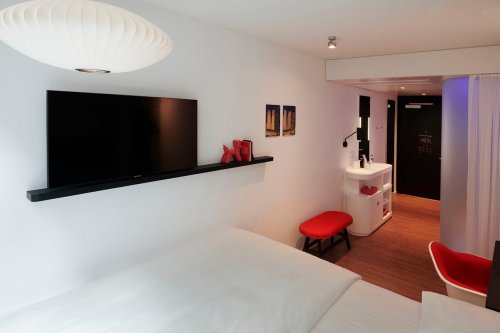 Modern hotel room featuring a bed, wall-mounted TV, red accents, and a sleek bathroom area.