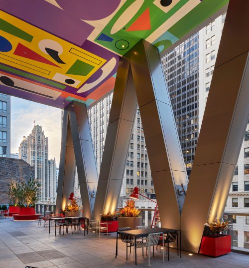 Modern outdoor space with colorful ceiling art, sleek pillars, and seating, surrounded by urban skyscrapers.