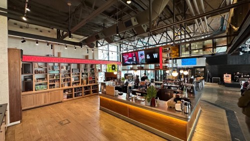 A modern café interior with a wooden counter, shelves of products, and large screens displaying menus and promotions.