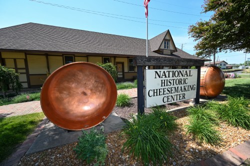 Sign for the National Historic Cheesemaking Center, flanked by large copper cheese molds and surrounded by greenery.