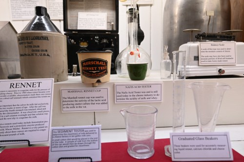 Display of historical cheese-making tools, including rennet testers, beakers, and a balance scale.