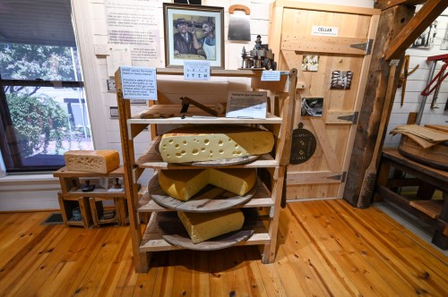 A wooden shelf displays various types of cheese in a museum setting, with informational signs and a door labeled "CELLAR."