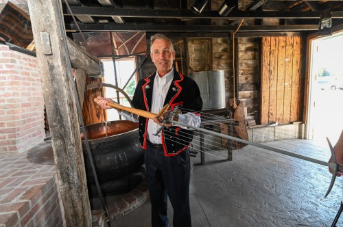 A man in a traditional outfit holds a large tool inside a rustic building with a copper pot in the background.