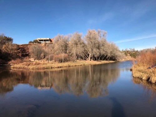 A serene landscape featuring a calm river, bare trees, and a house on a hill under a clear blue sky.