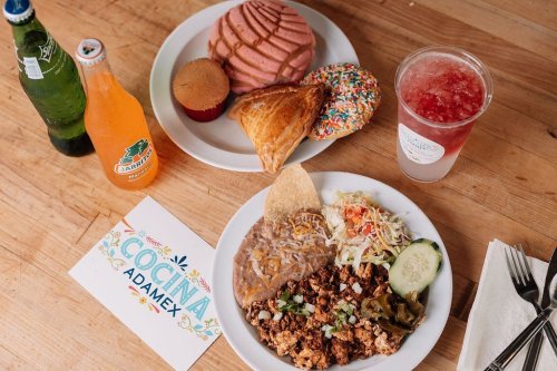 A colorful spread of Mexican food, pastries, and drinks on a wooden table, featuring tacos, sweets, and beverages.