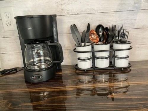 A coffee maker next to a utensil holder with various cutlery and kitchen tools on a wooden countertop.