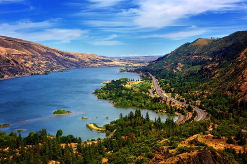 A scenic view of a river winding through green hills and mountains under a blue sky with scattered clouds.