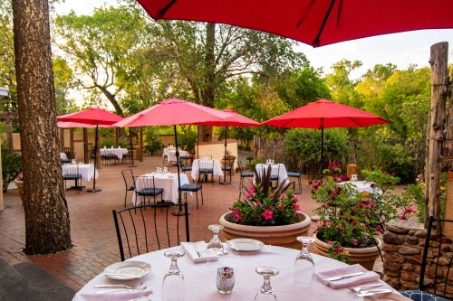 Outdoor dining area with red umbrellas, tables set for guests, surrounded by greenery and flowers.