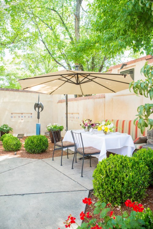 A serene outdoor dining area with a table under an umbrella, surrounded by greenery and colorful flowers.