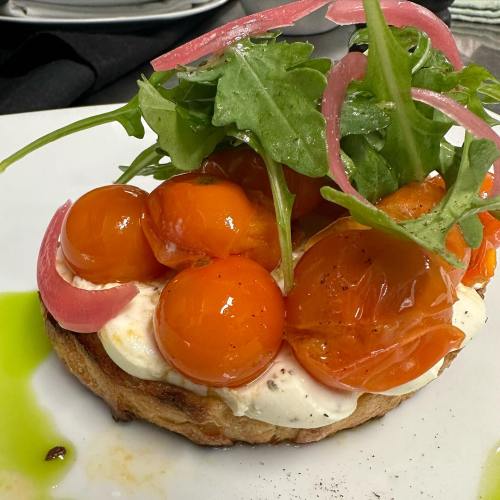 A toasted bread topped with creamy spread, vibrant orange tomatoes, pickled onions, and fresh arugula.
