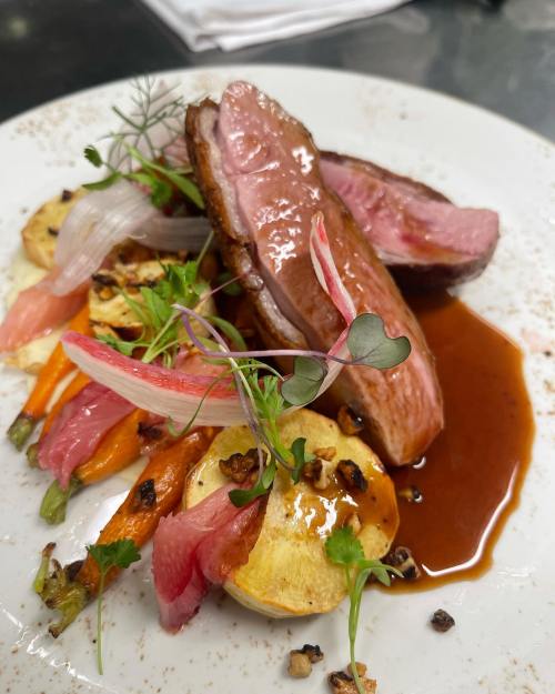A beautifully plated dish featuring sliced duck breast, colorful vegetables, and a rich sauce, garnished with microgreens.