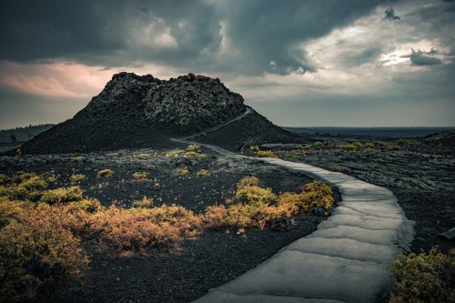 A winding path leads through a volcanic landscape with dark rock formations and patches of greenery under a cloudy sky.