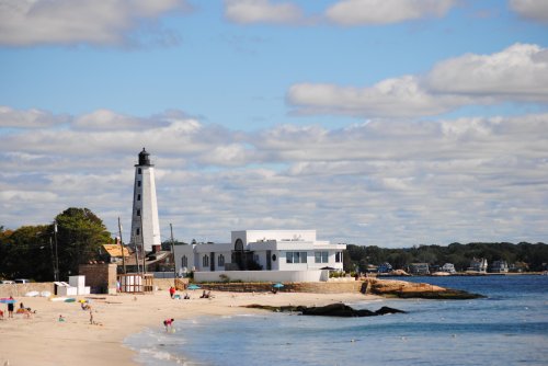 A scenic beach view featuring a lighthouse, white building, and cloudy blue sky. People enjoy the sandy shore.