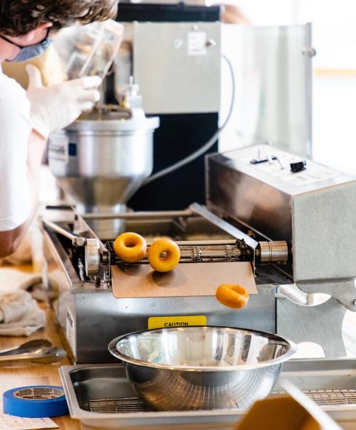 A person in gloves operates a donut machine, with freshly made donuts rolling off onto a conveyor.