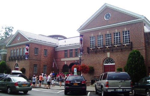 A brick building with large windows, people gathered outside, and decorative banners. Cars parked in front.