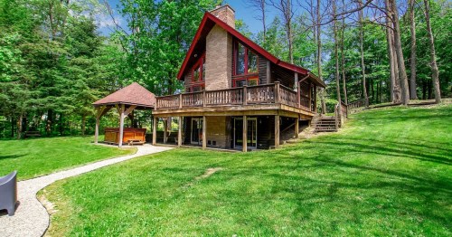 A charming wooden cabin with a red roof, surrounded by lush greenery and a pathway leading to a hot tub area.