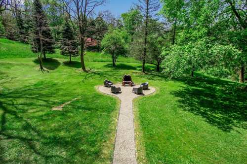 A serene outdoor space with a stone path leading to a fire pit surrounded by chairs, set in a lush green landscape.