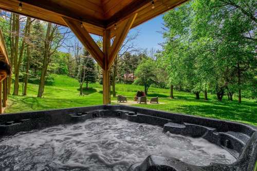 A hot tub under a wooden structure, surrounded by lush green trees and a sunny outdoor setting.