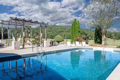 A serene outdoor pool surrounded by greenery, with lounge chairs and a pergola under a partly cloudy sky.