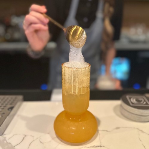 A bartender holds a gold spoon above a frothy cocktail in a decorative glass on a marble countertop.