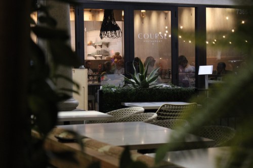 A cozy restaurant scene with indoor seating, plants, and warm lighting visible through large windows.