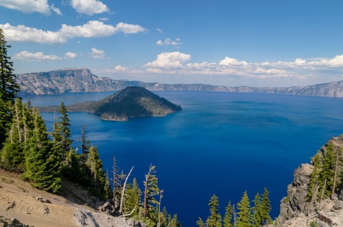 A panoramic view of Crater Lake, featuring deep blue water, a small island, and surrounding green trees under a clear sky.