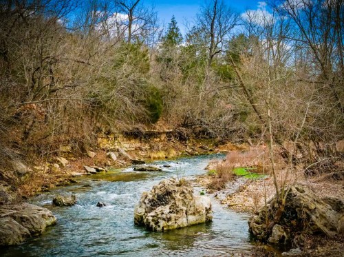 A serene river flows through a wooded area, surrounded by bare trees and rocky banks under a blue sky.