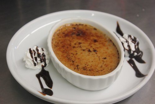 A creamy dessert in a white dish, topped with caramelized sugar, accompanied by whipped cream and chocolate drizzle.