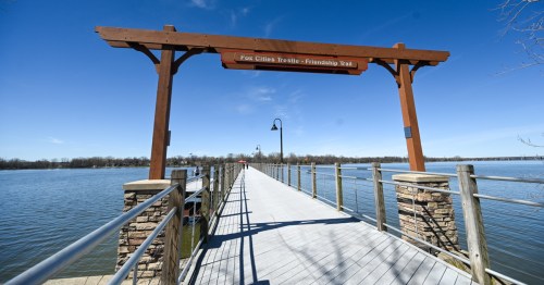 A wooden walkway leads over water, marked by a sign for the Fox Cities Trestle-Friendship Trail under a clear blue sky.