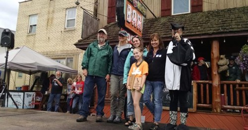 A group of six people stands on a stage at an outdoor event, smiling and posing for the camera.