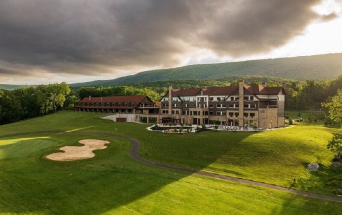 A scenic view of a hotel surrounded by green fields and mountains under a cloudy sky. A golf course is visible in the foreground.