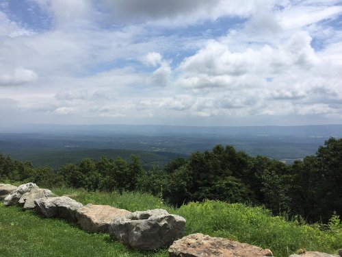 A scenic view from a mountain overlook, featuring rolling hills, a cloudy sky, and a rocky foreground.