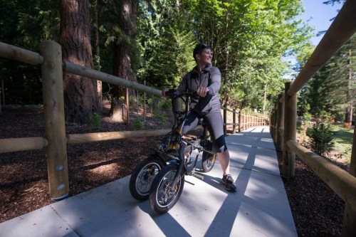 A person stands beside a small bike on a paved path surrounded by trees and wooden fencing.
